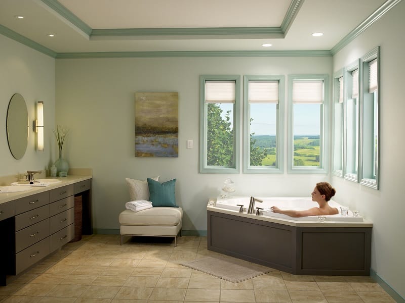 Woman looking out her windows as she lays in the bathtub and her cellular shades are half way down.