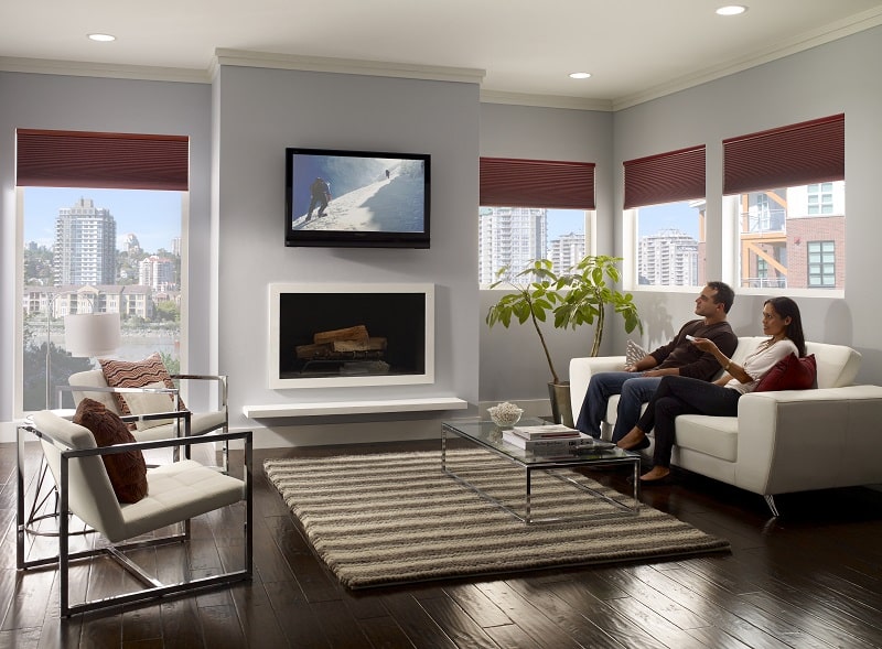 Husband and wife sitting on their couch watching television as the women lowers the motorized cellular shades with a remote.