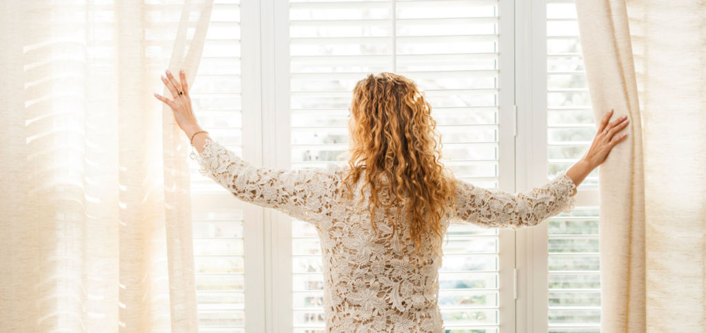 woman facing away, opening sheer curtains layered on top of white shutters