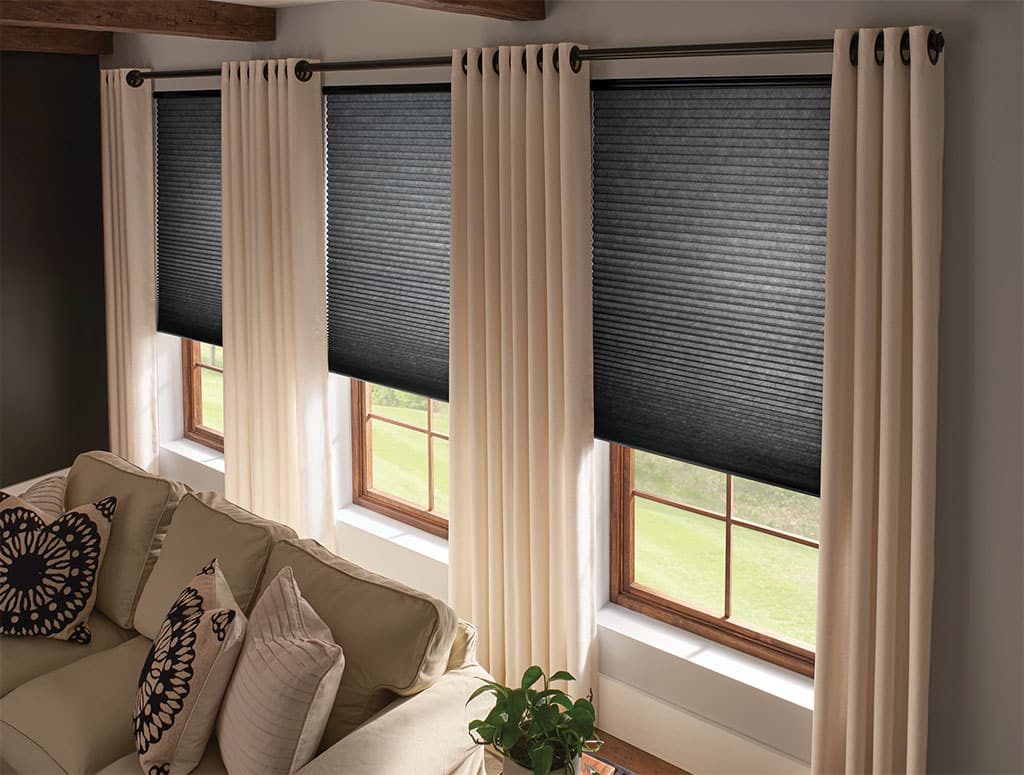 Living room with black cellular blackout shades layered behind beige grommet drapery panels on wood-framed windows for complete light and privacy control