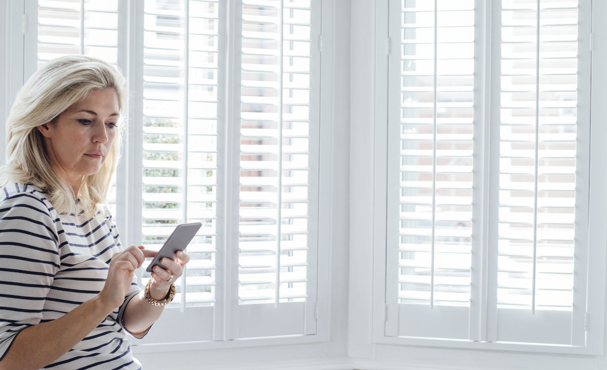 woman stands in front of modern white shutters on sunlight windows, controlling the motorized window coverings from her phone