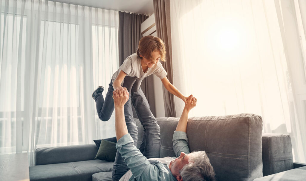 grandpa and grandson happy on couch with sheer curtains and grey drapes covering sunlit windows in the background