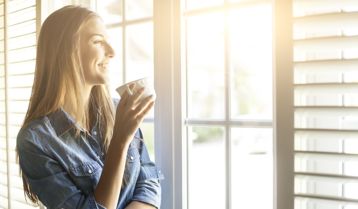 picture of a woman smiling and drinking tea looking out the window with wood shutters in a modern room